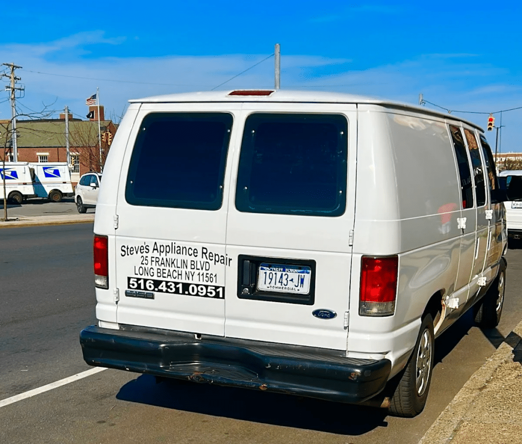 White appliance repair service van parked on a city street with business branding on the rear doors. Commercial service vehicle for “Steve’s Appliance Repair” displaying company address, Long Beach, NY location, and phone number “516.431.0951.” Appliance repair transportation and home service business concept featuring a Ford work van used for residential appliance maintenance, repair calls, and technician service visits.